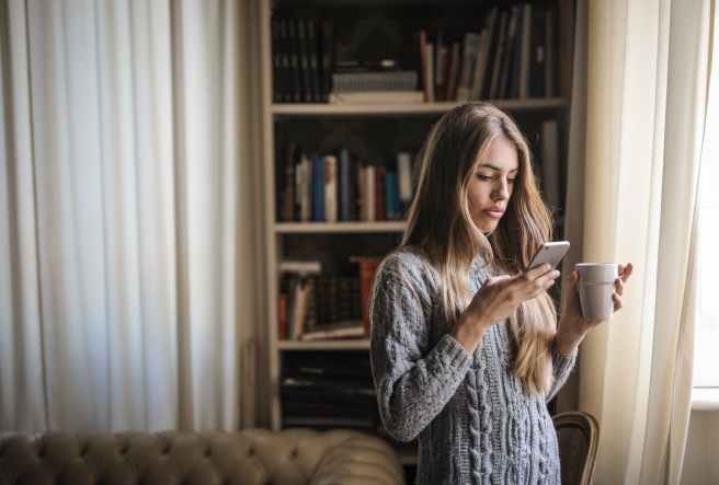 photo of woman in gray sweater holding white ceramic cup while using her phone